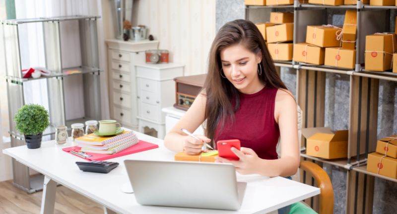 A imagem mostra uma mulher de cabelos castanhos soltos e uma blusa vermelha. Ela está sentada em uma mesa de escritório segurando um celular e olhando para um notebook. Atrás dela há uma estante com diversas caixas parecidas com caixas para entrega de produtos.