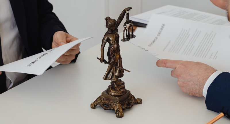 Dois homens fazem a leitura de um contrato de trabalho em cima de uma mesa, onde está posicionada uma miniatura da estátua da justiça (mulher vendada segurando uma balança).