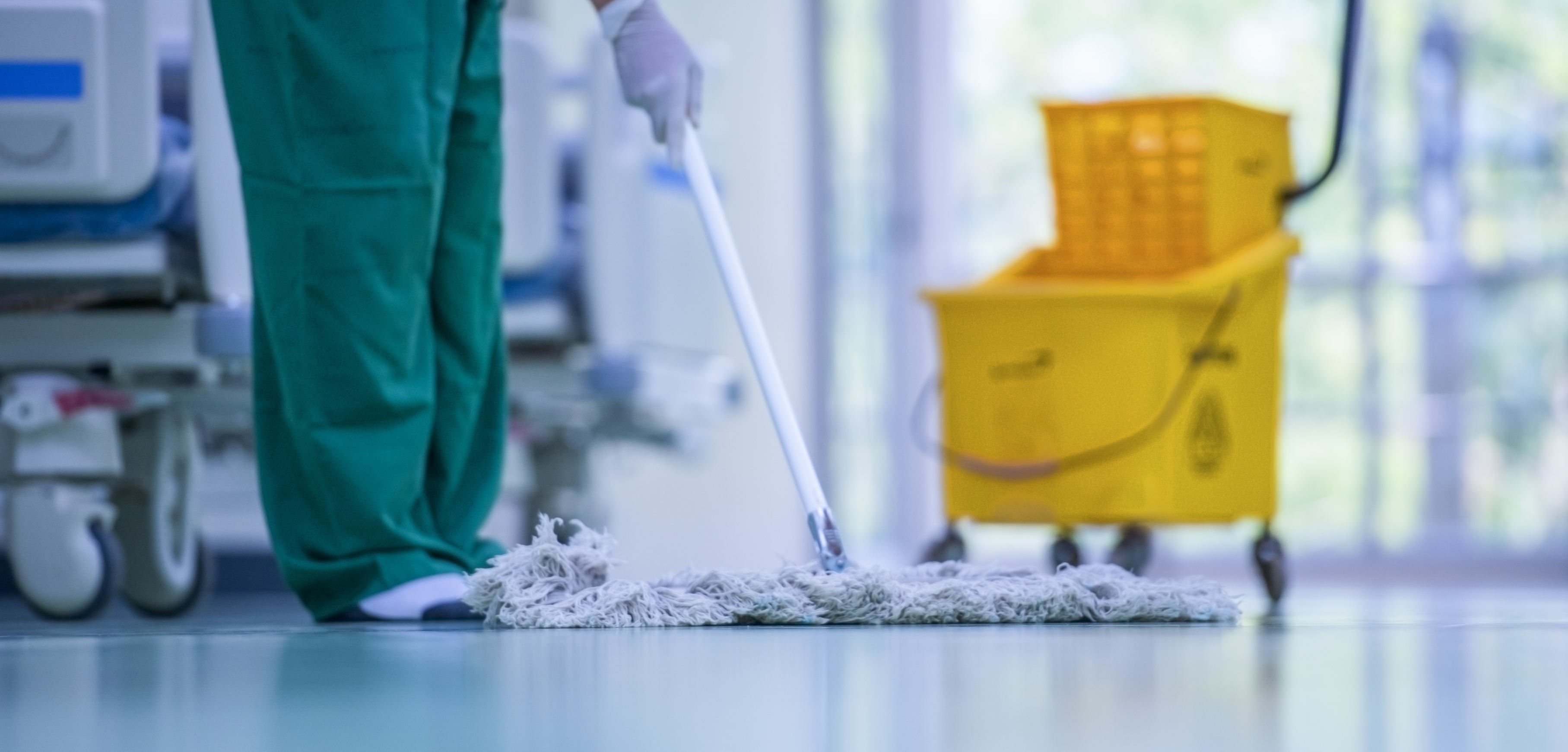 A imagem apresenta um trabalhador realizando a limpeza de um ambiente. Ele está vestido num uniforme verde e utiliza equipamentos de limpeza que costumam ser usados em ambiente hospitalar.