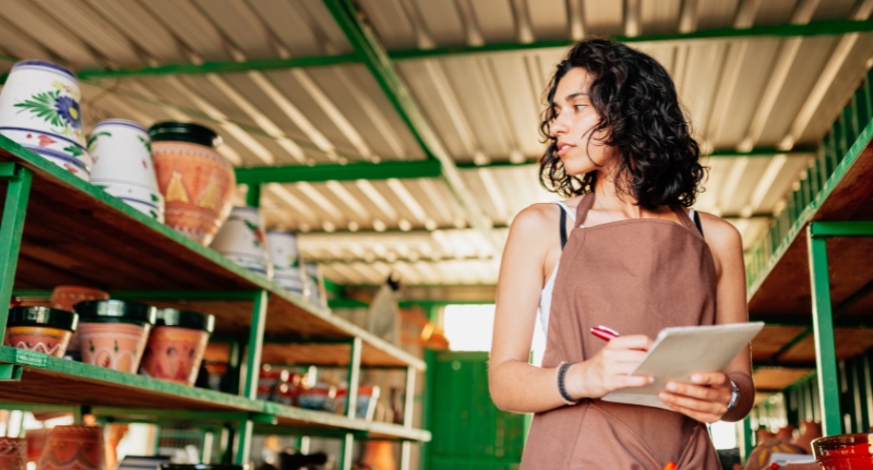 Mulher dentro de um estoque fazendo anotações enquanto observa prateleiras com vasos, representando o processo de controle e organização de inventário.