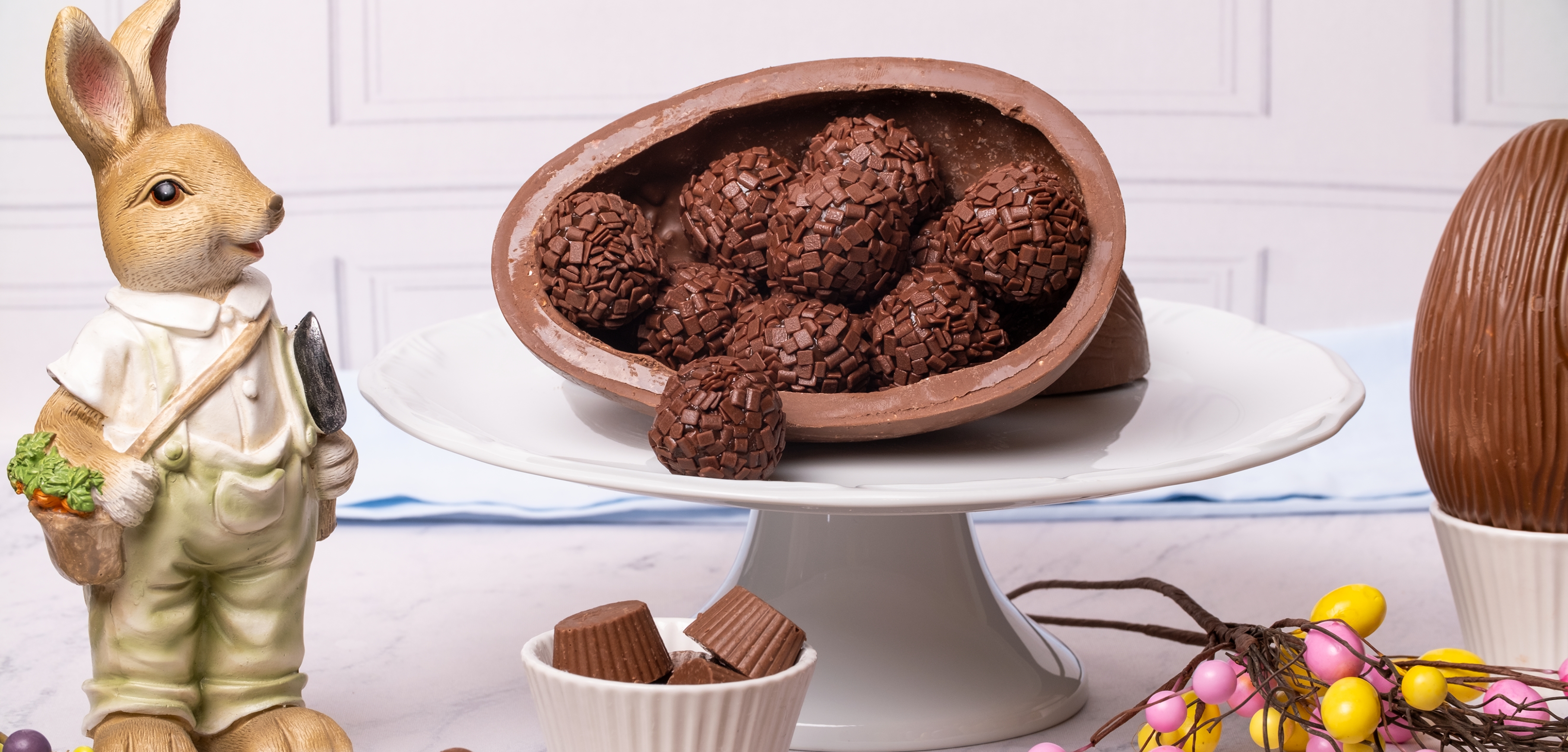 A imagem apresenta um ovo de páscoa com brigadeiros dentro em cima de um prato. Ao lado há algumas decorações, como um coelho, alguns bombons e ovinhos coloridos.