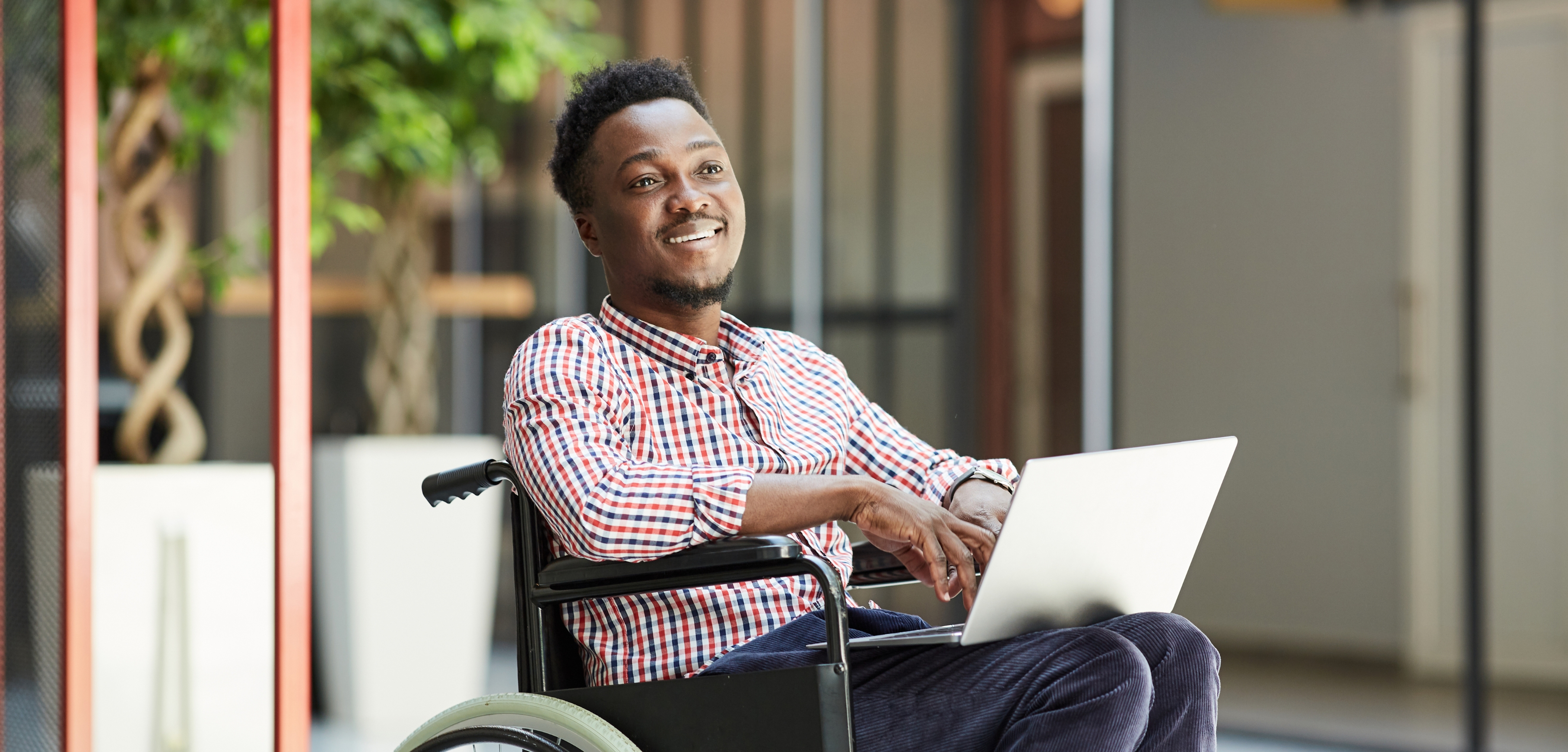 A imagem apresenta um homem negro, sentado em uma cadeira de rodas e segurando um laptop apoiado nas pernas. Ele veste uma camisa xadrez, calça escura e está sorrindo.