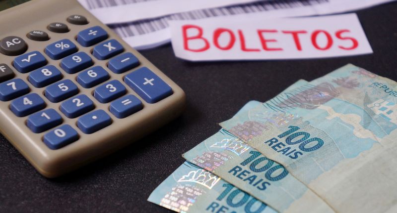 A imagem mostra uma calculadora com notas de 100 reais e um papel com a palavra "boletos".