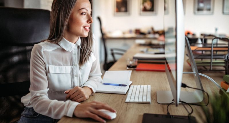 A imagem mostra uma mulher de cabelo liso castanho e camisa branca mexendo no computador.