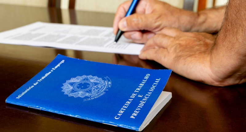 Um Carteira de Trabalho e Previdência está posta sobre uma mesa, enquanto uma pessoa assina seu contrato de trabalho.