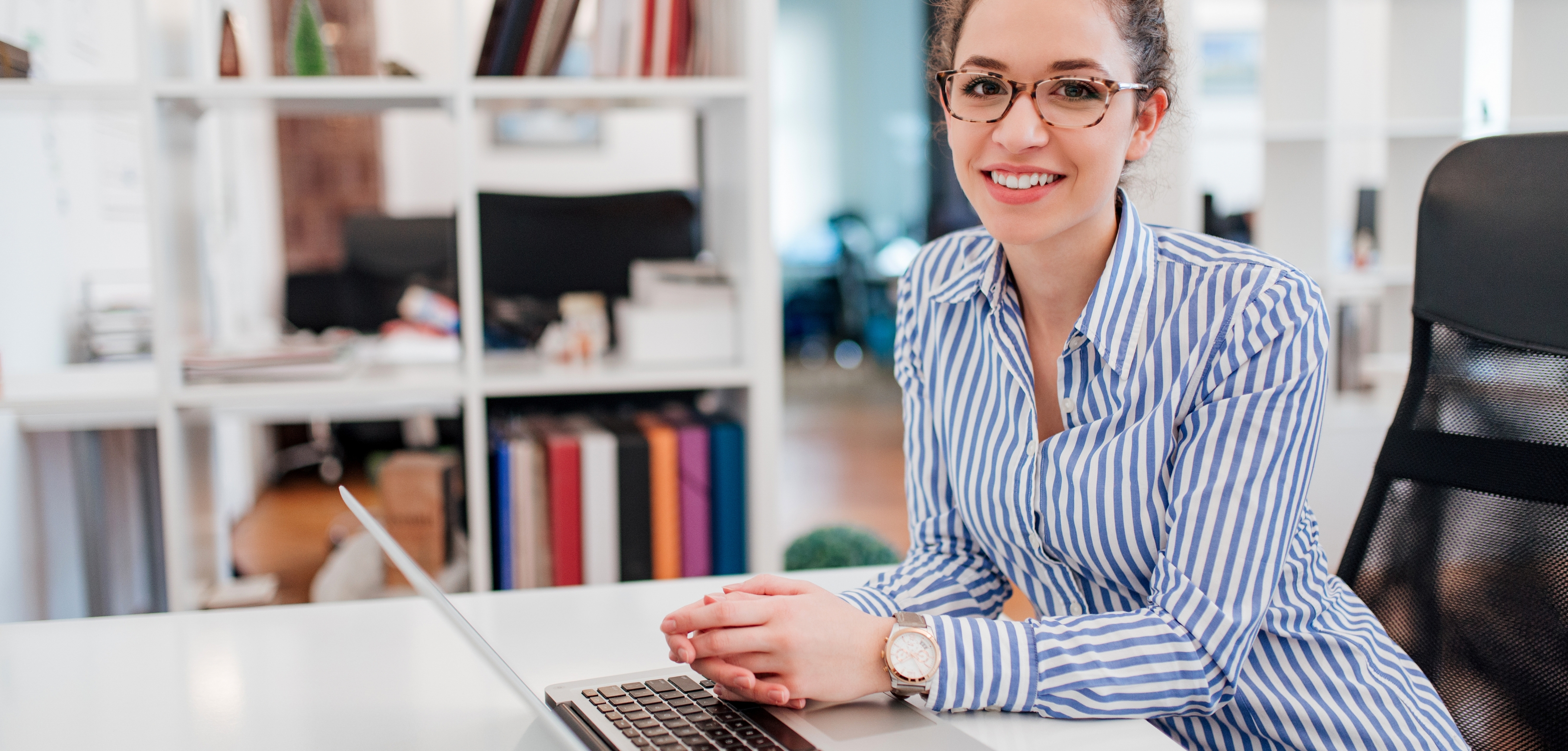 A imagem mostra uma mulher sorrindo, usando óculos e uma camisa listrada, sentada em uma mesa com um notebook. Ela está em um ambiente de escritório, onde ao fundo há uma estante com livros.