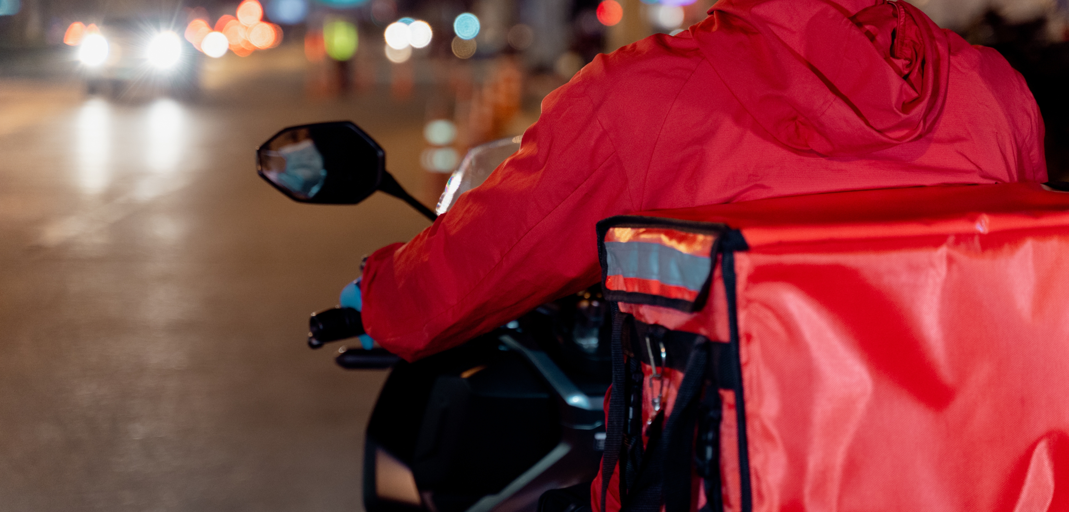 A imagem mostra as costas de um motoboy, vestido numa jaqueta vermelha e com uma bolsa de entregas também vermelha. Ele está pilotando em uma rua à noite.