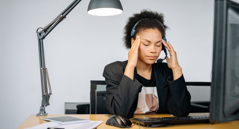 Profissional com headset sentada à mesa de trabalho, com as mãos nas têmporas, em referência à saúde mental no ambiente profissional.