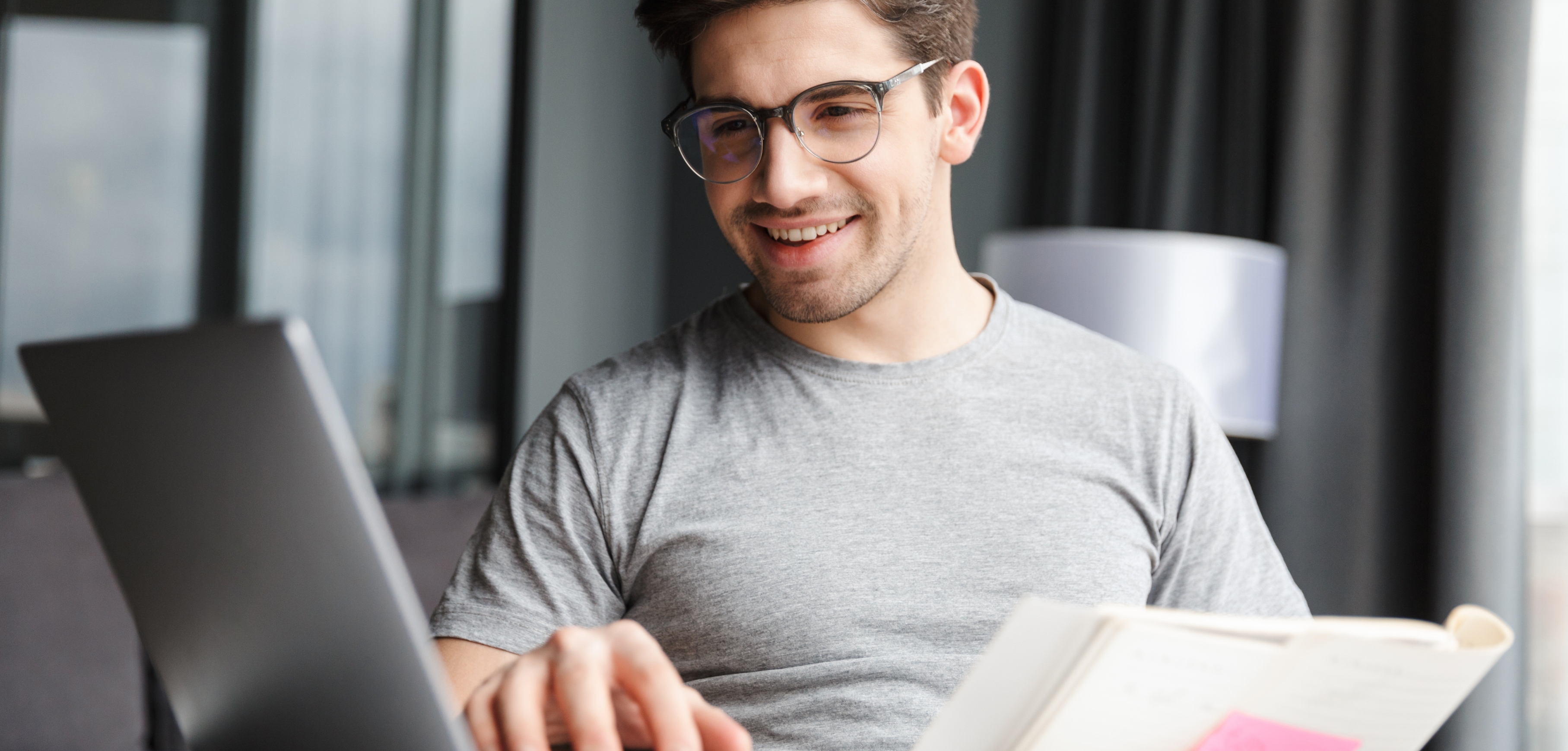 A imagem apresenta um homem branco, de cabelos castanhos e barba. Ele veste uma camiseta cinza e óculos de grau, está sentado segurando um caderno e olhando para uma tela de notebook.