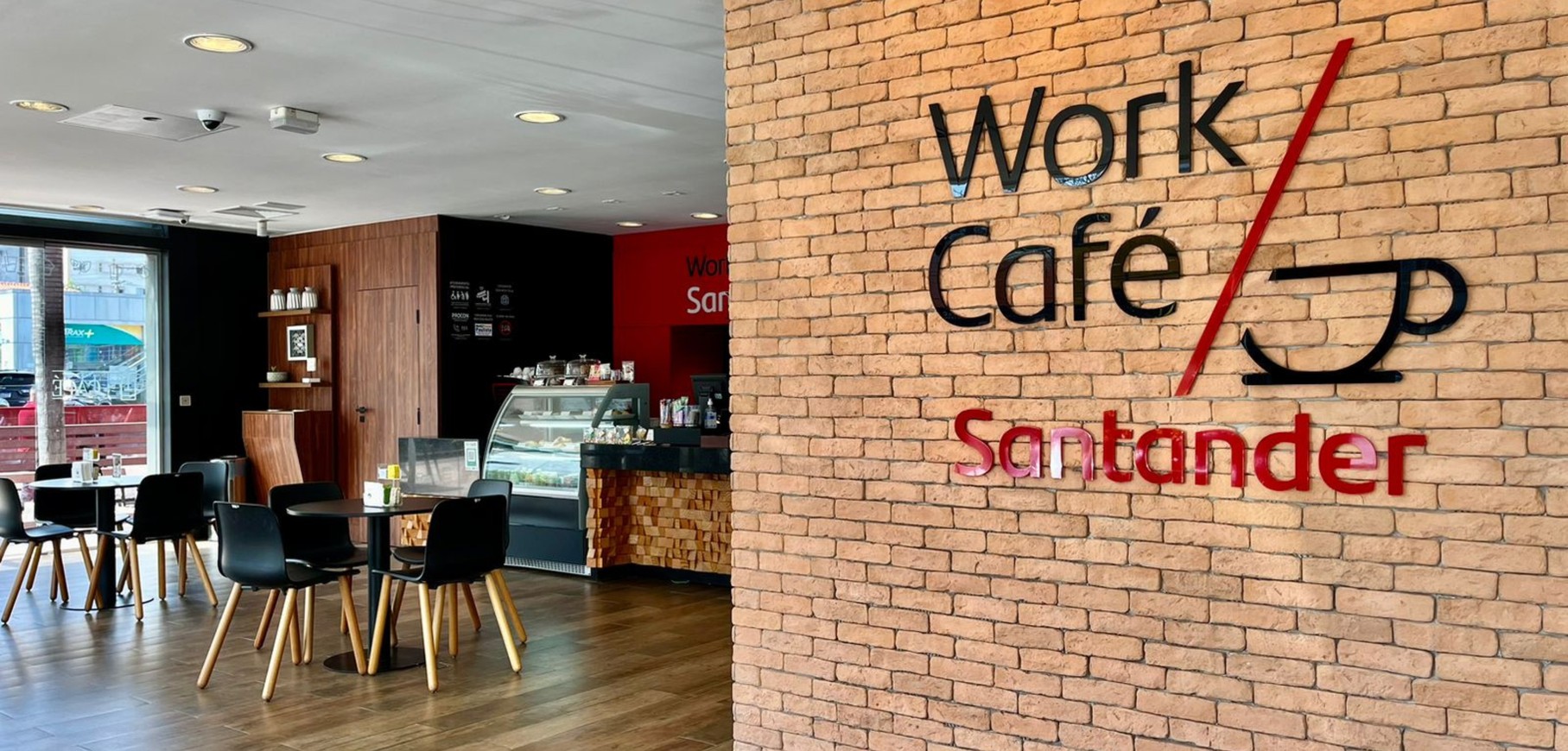 A imagem mostra um espaço Work/Café do Santander. Aqui se vê apenas o logo em uma parede de tijolos expostos.