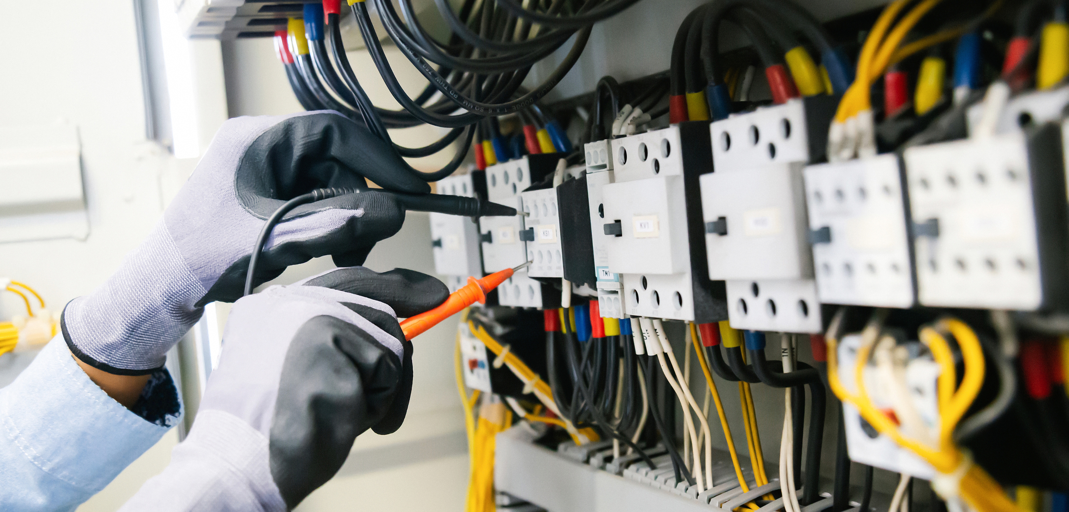 A imagem mostra duas mãos, com luvas de proteção, mexendo em um painel cheio de fios e componentes elétricos.