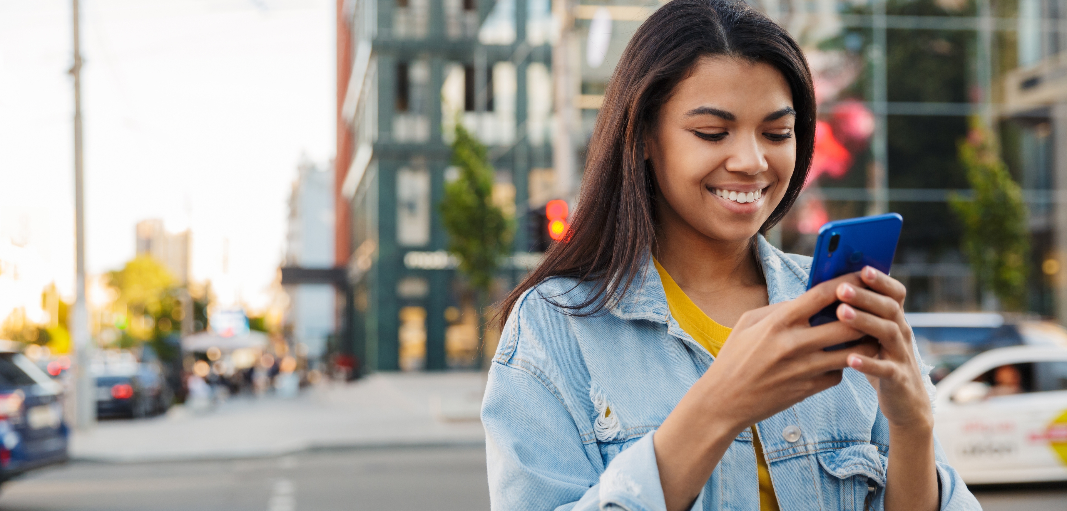 A imagem mostra uma mulher jovem, de cabelos castanhos e pele morena, sorrindo enquanto utiliza um celular azul. No fundo da imagem, desfocado, há uma rua, com prédios e árvores ao fundo.