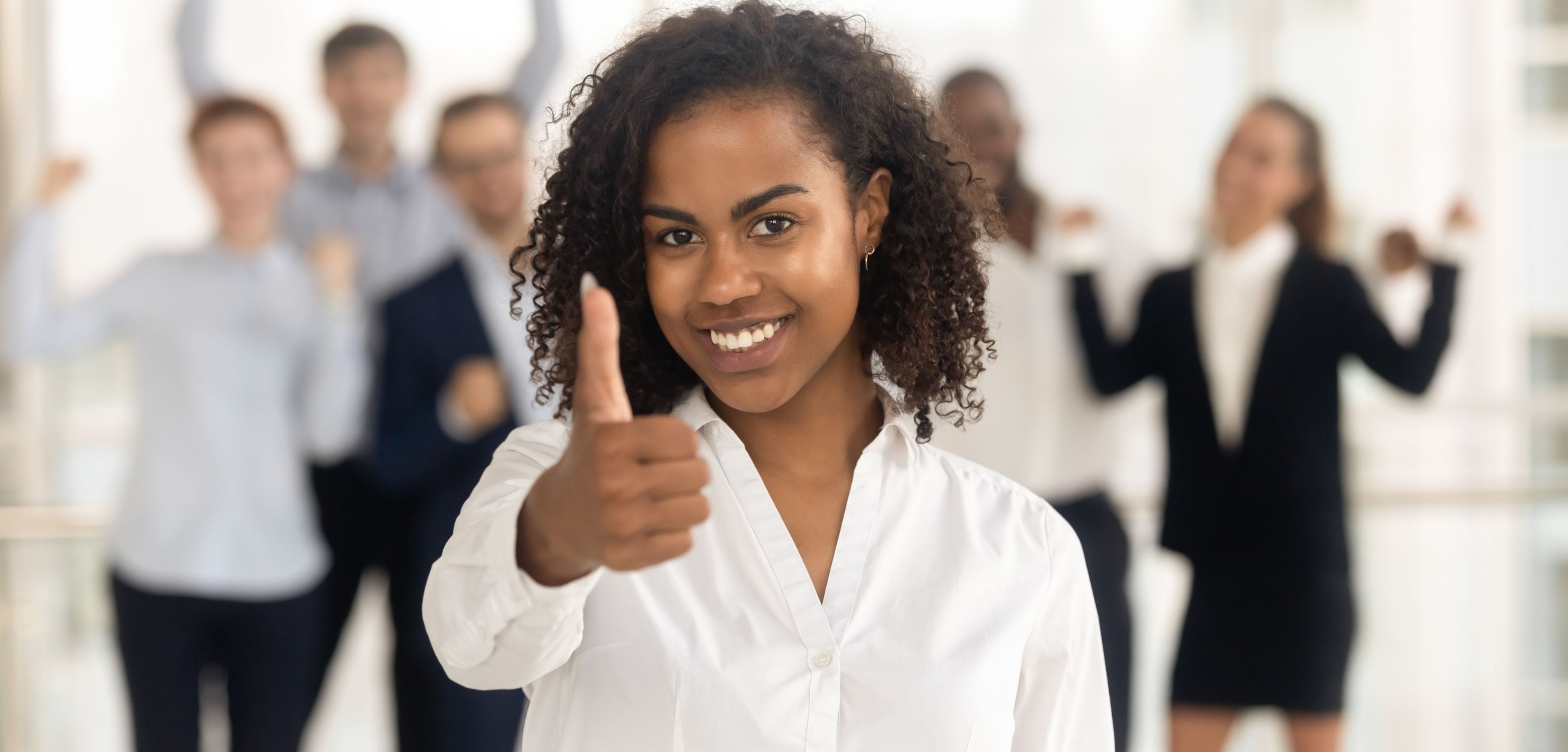 A imagem mostra uma profissional fazendo um sinal de "joia" com a mão, para expressar algo positivo. Ela está vestidacom uma camisa branca social e atrás dela, desfocados, existem outras pessoas de roupa social comemorando.