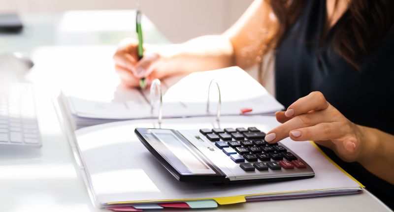 Uma mão feminina fazendo contas do faturamento do seu MEI com auxílio de uma calculadora e papéis.